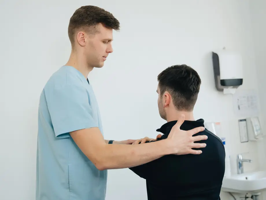 A physiotherapist stands behind a seated patient and checks the movement of his shoulder.