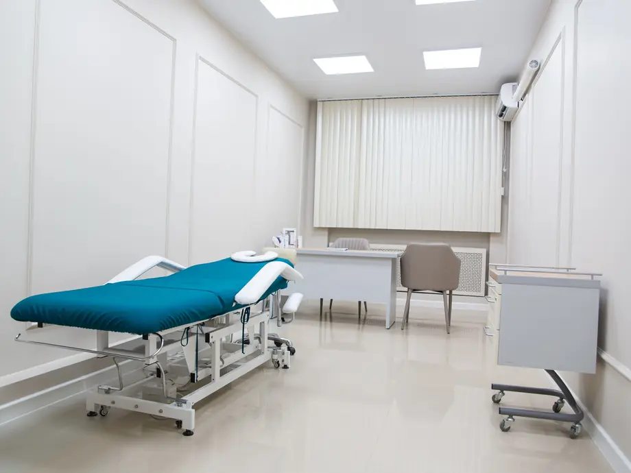 An empty treatment room with a padded couch, a desk and two chairs, and blinds at the window.