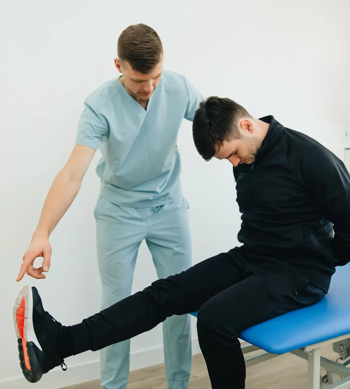 A physiotherapist guides a seated patient through a leg stretch on a treatment couch in a bright clinic room.