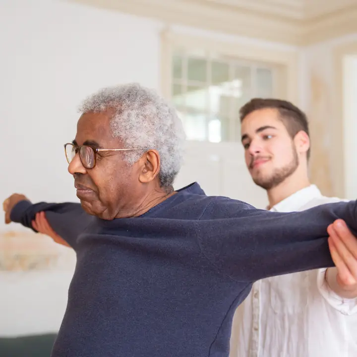 An older man stretches both arms out to the sides while a therapist supports them.
