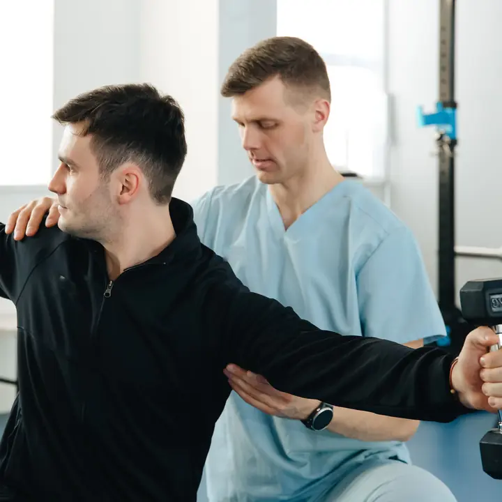 A patient raises a small dumbbell while a physiotherapist supports his arm and shoulder.